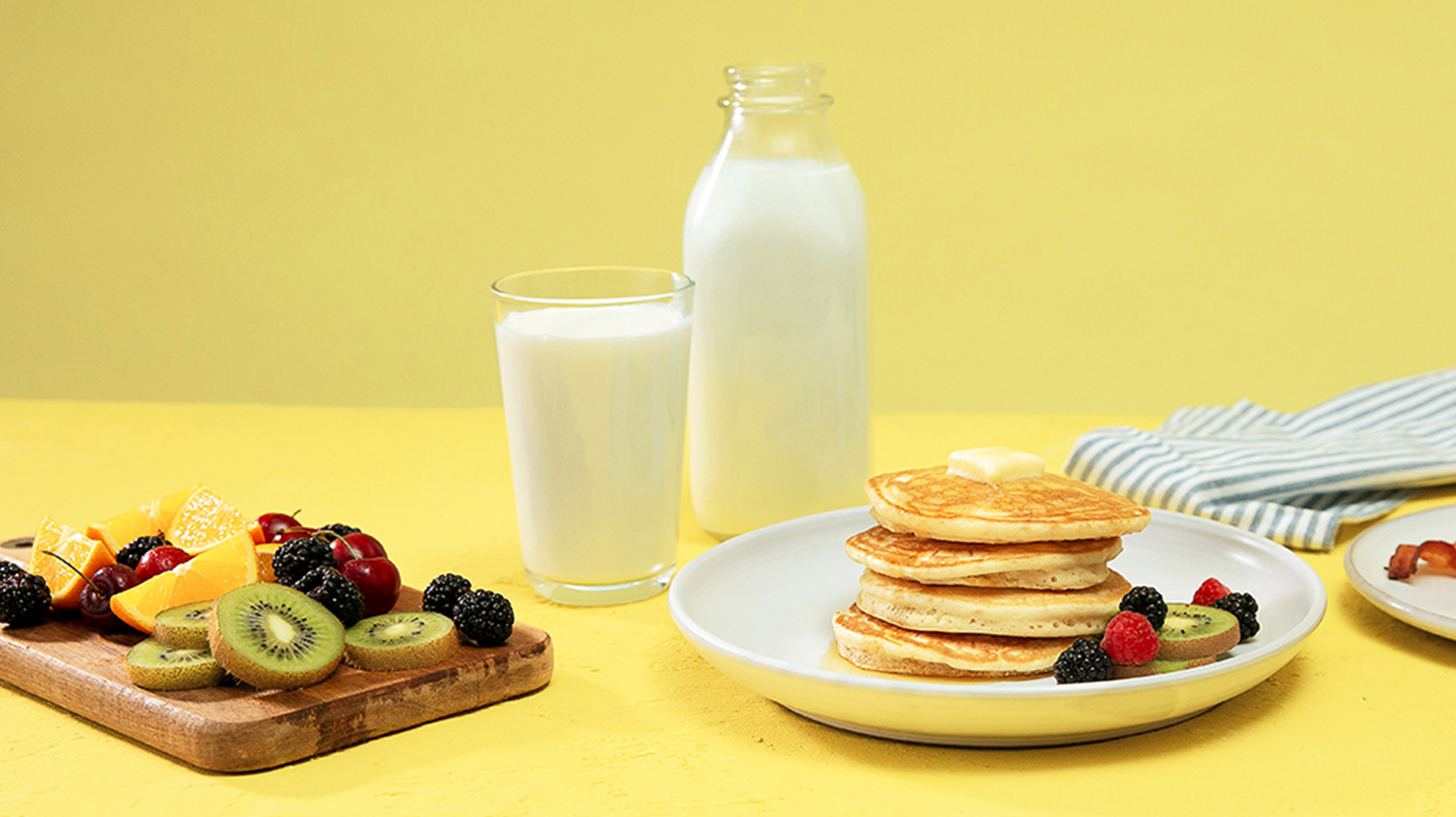 Stack of pancakes with a big glass of milk and a plate of fruit.