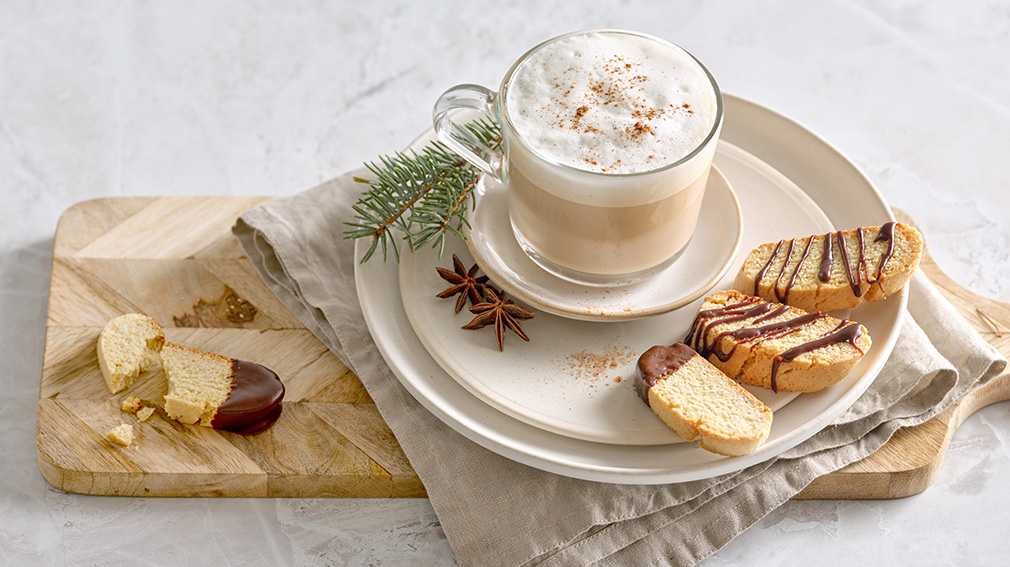 A round white plate with biscotti cookies with chocolate drizzle on top and a cappuccino in a small clear mug.