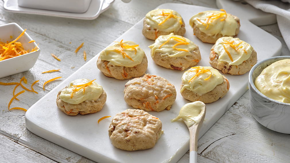 Orange Carrot cookies with orange cream cheese frosting on white serving board with white spoon.