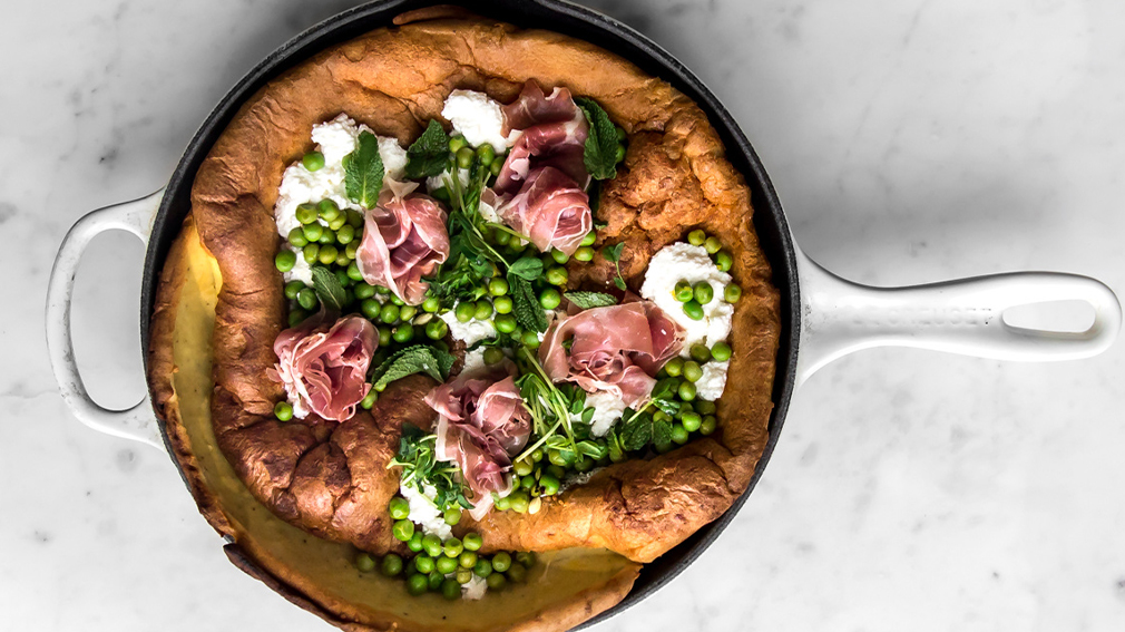 Overhead shot of white cast iron pan with dutch baby with peas, ricotta, prosciutto and herbs on top.