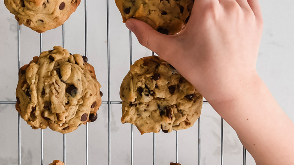 Overhead shot of a childs hand grabbing a chocolate chip cookie off of a cooling rack with other cookies.