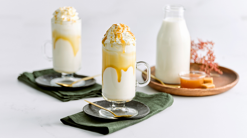 Two tall, clear glass mugs filled with steamed milk topped with whipped cream and drizzled butterscotch sauce, on small round black plates on top of dark green napkins. 