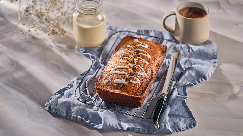 A loaf of eggnog banana bread with a white icing drizzle on top resting on a metal cooling rack on top of a light blue napkin next to a black handled bread knife.