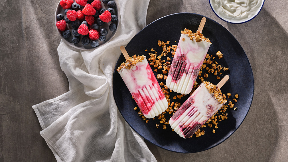 Overhead view of three frozen morning yogurt fruit popsicles on a bed of granola on a round black plate next to a round dark bowl full of raspberries and blackberries.