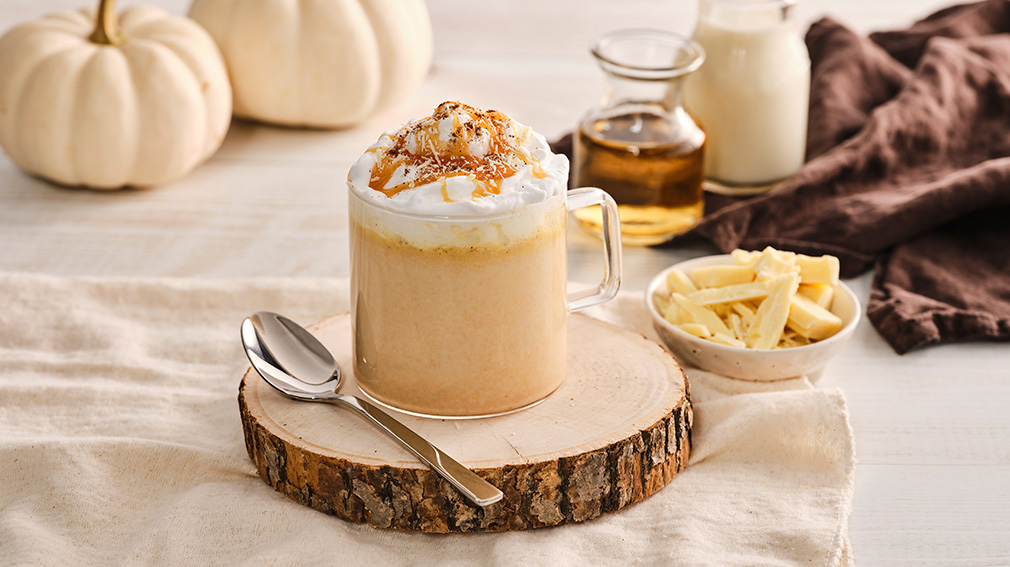 Clear glass mug with pumpkin spice white hot chocolate topped with a dollop of whipped cream and drizzle of caramel sauce sitting on top of a small, round, wooden serving board next to a metal spoon.