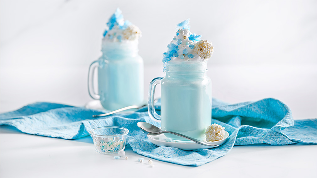 Two large clear glass mugs with blue hot chocolate topped with whipped cream dollop and white and blue sprinkles next to a blue napkin, spoon, and a small white bowl containing sprinkles.